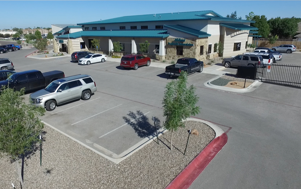 Exterior view of medical clinic building. Steel frame supports white stucco and rock wall facade with a turquoise metal roof. Parking lot with assorted cars and tidy landscaping
